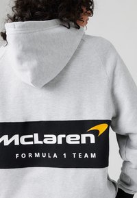 Szary hoodie z czarnym poziomym pasem z napisem "McLaren" i "Formula 1 Team" w białym kolorze. Posiada kaptur i miękką teksturę.