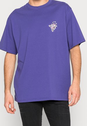 Mężczyzna ubrany w fioletowy T-shirt z krótkim rękawem z małą kwiatową grafiką i napisem na lewej piersi, połączony z czarnymi spodniami.