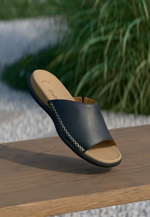 Black leather open-toe slide sandal with beige cushioned insole and decorative white cross-stitching on wooden surface against green plants.
