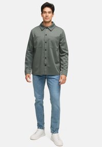 Olivengrøn jakke med knaplukning foran, to brystlommer og spredt krave, parret med lyseblå jeans og hvide sneakers.