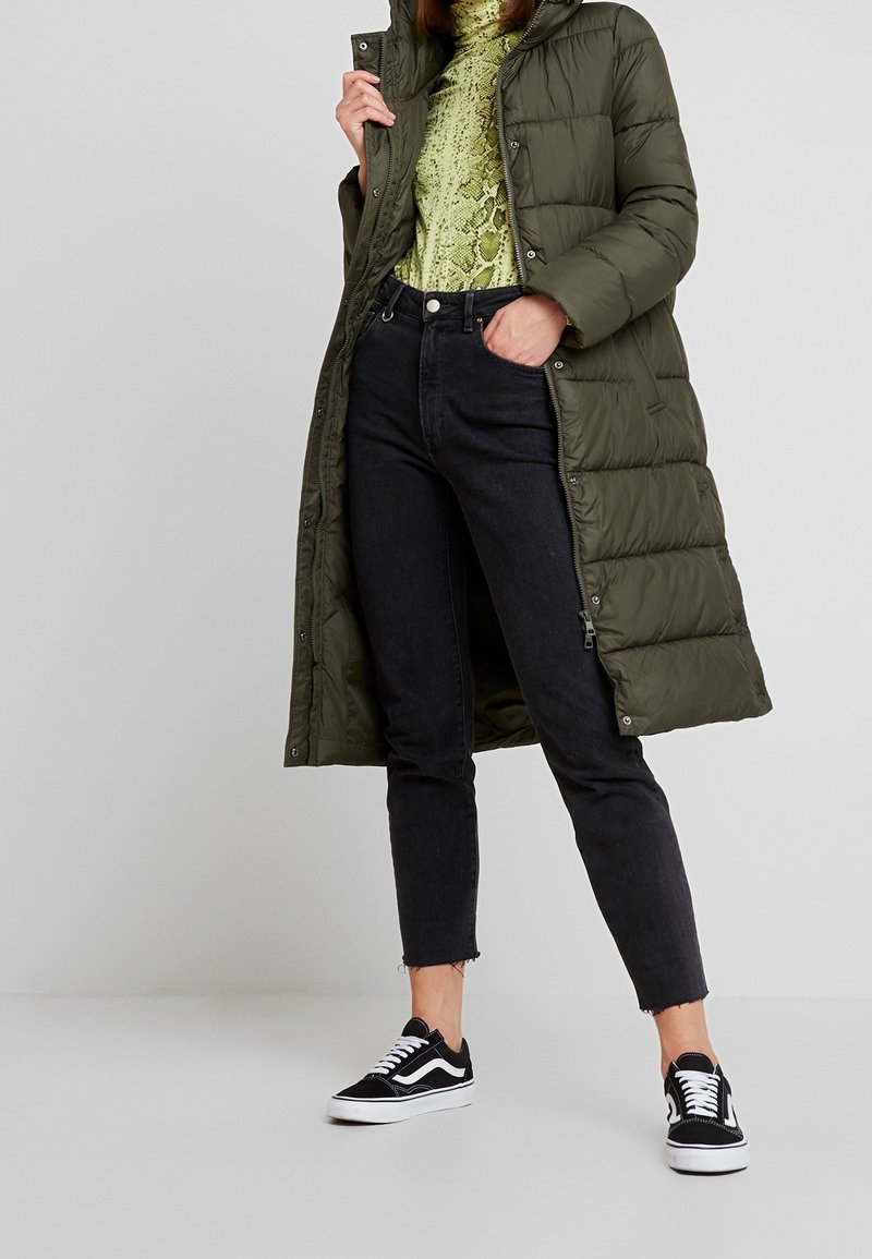 Femme portant un long manteau matelassé vert olive, un haut vert imprimé serpent, un jean noir court et des baskets noires et blanches, debout avec une main dans la poche.