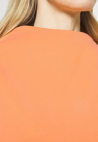 Lyhythihainen, pyöreäkauluksinen paita kirkkaan oranssina, valmistettu pehmeästä, kevyestä kankaasta, joka tuntuu sileältä ja jossa on minimalistiisia yksityiskohtia.