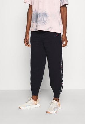 Osoba ubrana w czarne spodnie dresowe z logo Tommy Hilfiger po boku, jasną koszulkę w stylu tie-dye oraz białe trampki z brzoskwiniowymi akcentami.