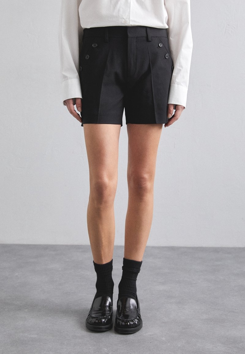 Shorts noirs sur mesure avec double boutonnerie et plis avant, fabriqués dans un tissu lisse. Portés avec des chaussettes noires et des mocassins brillants.