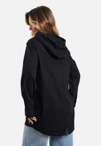 Veste noire à capuche avec des manches longues, une taille cintrée et une fente à l'arrière. Fabriquée en tissu en coton, elle présente une coupe décontractée et un minimalisme dans ses détails.