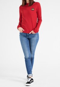 Femme portant une chemise à manches longues rouge Vans, un jean skinny bleu et des baskets noires Vans, se tenant les mains dans les poches sur un fond blanc.