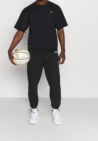 Hombre vestido con un chándal negro de Nike y zapatillas blancas, sosteniendo un balón de baloncesto blanco y dorado de Nike contra un fondo liso.
