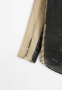Vêtement à manches longues avec motif tie-dye noir et beige, poignets boutonnés et coutures visibles sur fond clair.