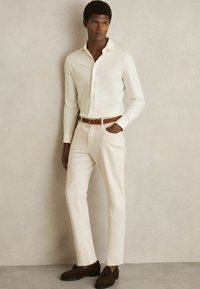 Cămașă albă cu mâneci lungi și guler butonat, asociată cu pantaloni de culoare deschisă și pantofi din suede închis. Un curea din piele maro completează ținuta.