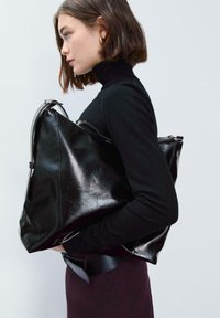 Borsa in pelle nera con forma triangolare, texture lucida e dettaglio della cerniera argentata, tenuta sotto il braccio da una persona con una maglietta nera.