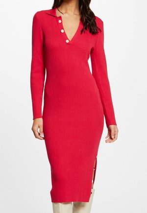 Długa czerwona sukienka z prążkowanego dzianego materiału, z długimi rękawami, dekoltem w serek z trzema guzikami oraz bocznym rozcięciem z detalami na guziki, noszona z białymi kozakami.