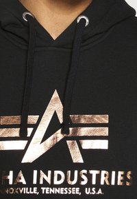 Sudadera negra con capucha que presenta un logo metálico en oro rosa con formas geométricas. Incluye cordones ajustables y ojales. Textura suave.