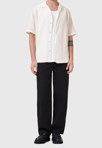 Homme portant une chemise blanche à manches courtes boutonnée par-dessus un sous-vêtement blanc, un pantalon noir et des chaussures noires, avec un tatouage de brassard noir sur le bras gauche.