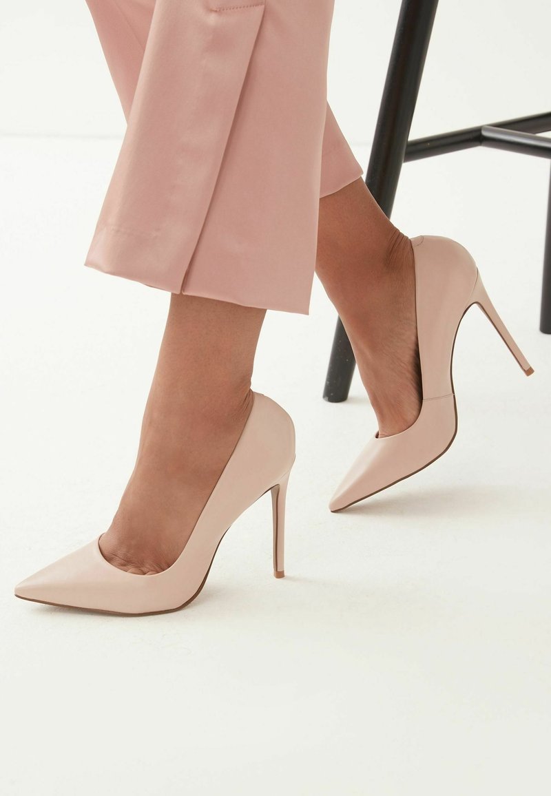 Femme portant des chaussures à talons hauts pointues rose blush assorties à un pantalon raccourci, assise près de pieds de chaise noirs.