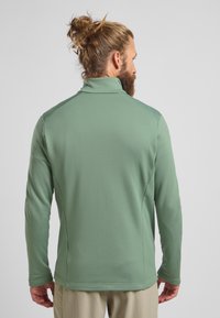 Pull en maille vert à manches longues, avec un col montant et des coutures plates, présenté de dos avec des poignets ajustables.