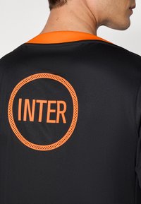 Svart sportströja med en orange cirkulär logotyp och texten "INTER" i mitten. Slät tygkvalitet och en kontrasterande orange krage.