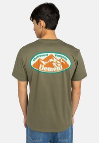 Olive zelené bavlnené tričko so širokým oválnym grafickým motívom na chrbte s horami a názvom značky v oranžovej a tealovej farbe. Pohodlný strih, krátke rukávy.