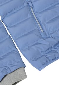 Doudoune bleu clair avec un motif matelassé horizontal, fermeture éclair argentée et bord de sweat-shirt gris dépassant de l'ourlet inférieur.