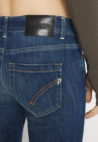 Pantalones vaqueros de mezclilla en azul oscuro con un parche de cuero de la marca en la parte trasera, con costuras naranjas y un acento plateado en el bolsillo trasero.