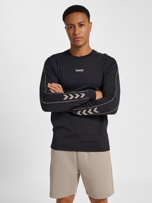 Man draagt een zwart shirt met lange mouwen met chevronpatronen op de mouwen en beige korte broek, staand met gekruiste armen tegen een witte achtergrond.