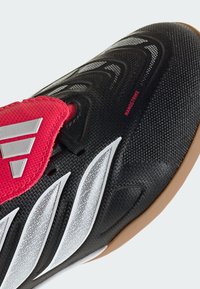 Chaussure de soccer d'intérieur noire avec des bandes blanches, une languette rouge portant un logo argenté, et une semelle en gomme, étiquetée "NANOSTRIKE" sur la tige.