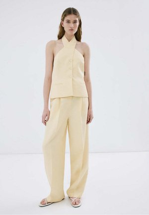 Kobieta w bladożółtym topie bez rękawów na szyję z guzikami, w połączeniu z szerokimi spodniami i białymi sandałami, stoi na jednolitym tle.