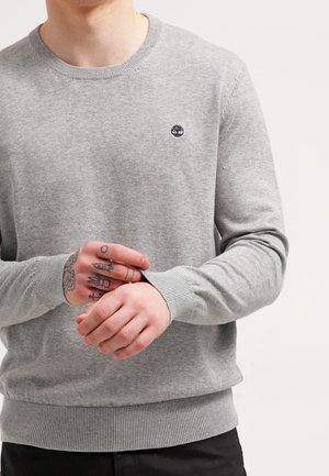 Osoba w jasnoszarym swetrze typu crewneck z małym czarnym logo drzewa, pokazująca wytatuowane palce podczas regulowania rękawa.