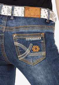 Dunkelblaue Denim-Jeans mit kontrastierendem orangefarbenem Nähten. Mit einer Gesäßtasche mit weißem Garnmuster und gebrandetem Lederpatch. Silberner Nieten-Gürtel.