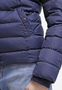 Námorně modrá prošívaná bunda se zapínáním na cvoky a žebrovanými manžetami, nošená přes světle modré džíny.