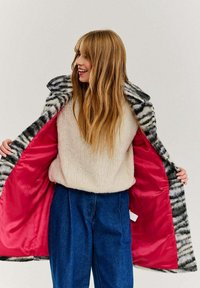 Umelá kožušinová bunda s tigrovými pruhmi v čierno-bielej farbe, s ružovou saténovou podšívkou, nosená cez béžový sveter a modré široké nohavice.