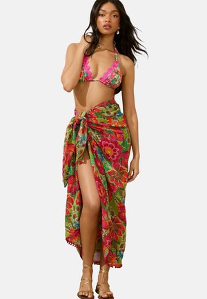 Bikini with scalloped edges in bright pink and a floral sarong featuring multicolored flowers on a green background. Gold sandals are worn.