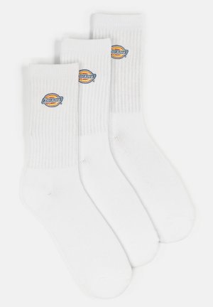 Trois paires de chaussettes blanches à crew avec des poignets côtelés. Chaque chaussette arbore un logo "Dickies" circulaire en bleu, jaune et rouge. Fabriquées en mélange de coton.