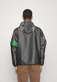 Man wearing shiny dark gray hooded jacket with green stripes on left sleeve, facing away, paired with brown pants.