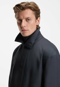 Cappotto nero, impermeabile, con colletto alto, caratterizzato da un design elegante e una texture liscia, esposto su uno sfondo neutro.