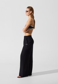 Pantalon large à taille élastique noire avec une texture lisse. Le modèle porte un top court contrasté noir et blanc ainsi que des lunettes de soleil.