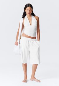 White halter top paired with white drawstring shorts. Material appears lightweight; model carries a matching white bag, wearing flip-flops.
