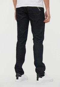 Jeans de mezclilla de un azul oscuro con corte slim, con costuras amarillas en contraste y dos bolsillos traseros, combinados con zapatillas blancas.