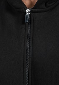 Primo piano di una felpa nera con zip, con una cerniera argento visibile e un tirante nero, indossata parzialmente chiusa vicino al collo.