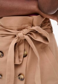Jupe beige cintrée avec une large ceinture, dotée d'une ceinture à nouer et de grands boutons marron. Le tissu semble lisse avec un design structuré.