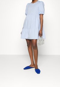 Světle modré šaty s nadýchanými rukávy a vrstveným lemem, vyrobené z bavlny. V kombinaci s modrými balerínami a průhlednou kabelkou, která zdůrazňuje jasné designové prvky.