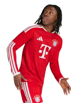Jonge man met dreadlocks die een rood FC Bayern München voetbaltenue draagt, omhoog kijkend met licht gespreide armen.