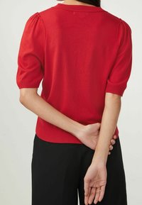 Pull rouge à manches courtes avec ourlet côtelé et manches bouffantes. Texture lisse, design ajusté avec un col rond. Porté avec un pantalon noir.