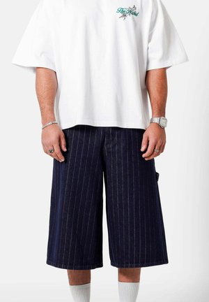 Mand iført hvid oversized t-shirt og mørkeblå stribede vidde shorts, med sølvur og ringe på hænderne.
