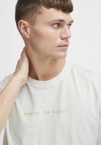 Lys beige bomull T-skjorte med rund hals, med gult brodert tekst "HVEM HAR HASSE?" på forsiden. Glatt tekstur.
