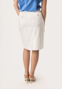 Witte penseelrok met een achterzijde-split, voorzien van twee achterzakken en zilveren knopen. Draagt met beige hakken en een blauwe top. Gladde stofstructuur.