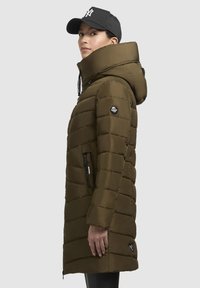 Manteau rembourré vert olive avec un col haut et une capuche amovible. Présente un design matelassé, des poches latérales zippées et un patch logo sur la manche.