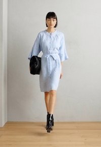 Svetlo modra lanena obleka s cinčnim pasom, kratkimi zvončastimi rokavi in gumbom spredaj. V kombinaciji z velikim črnim ženskim nahrbtnikom in črnimi čevlji z višino.