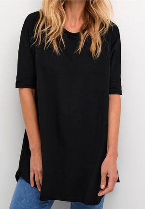 Femme blonde portant un t-shirt noir à manches courtes ample et un jean bleu, debout contre un mur blanc uni.