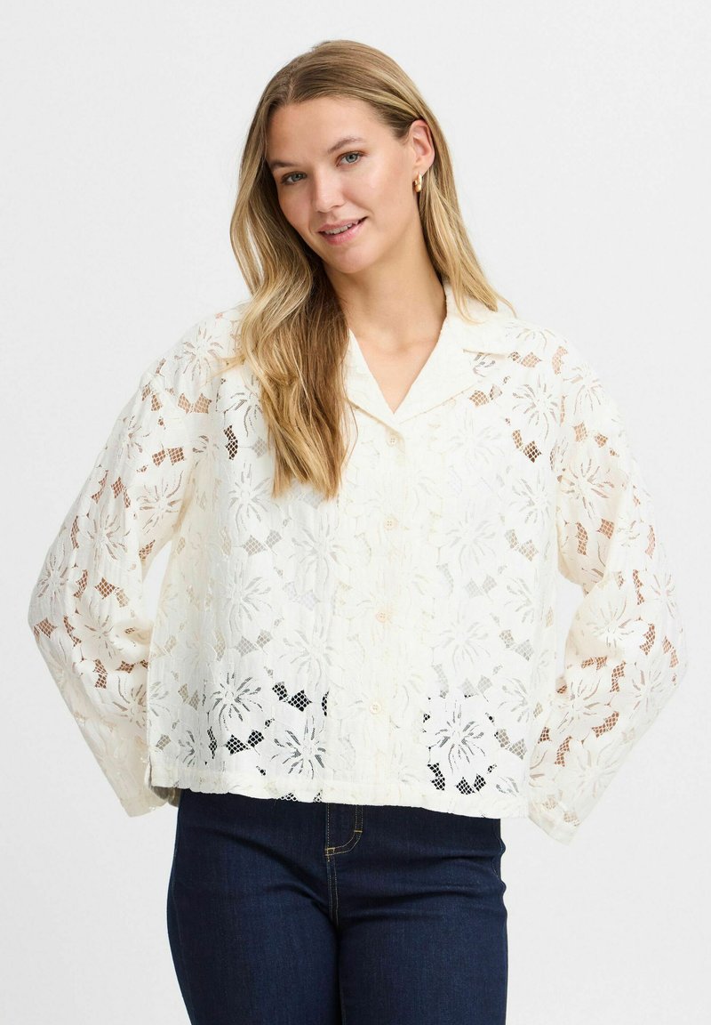 Witte kanten blouse met bloemenpatronen, knoopsluiting aan de voorkant, kraag en losse, lange mouwen. Draagt over donkere jeans.