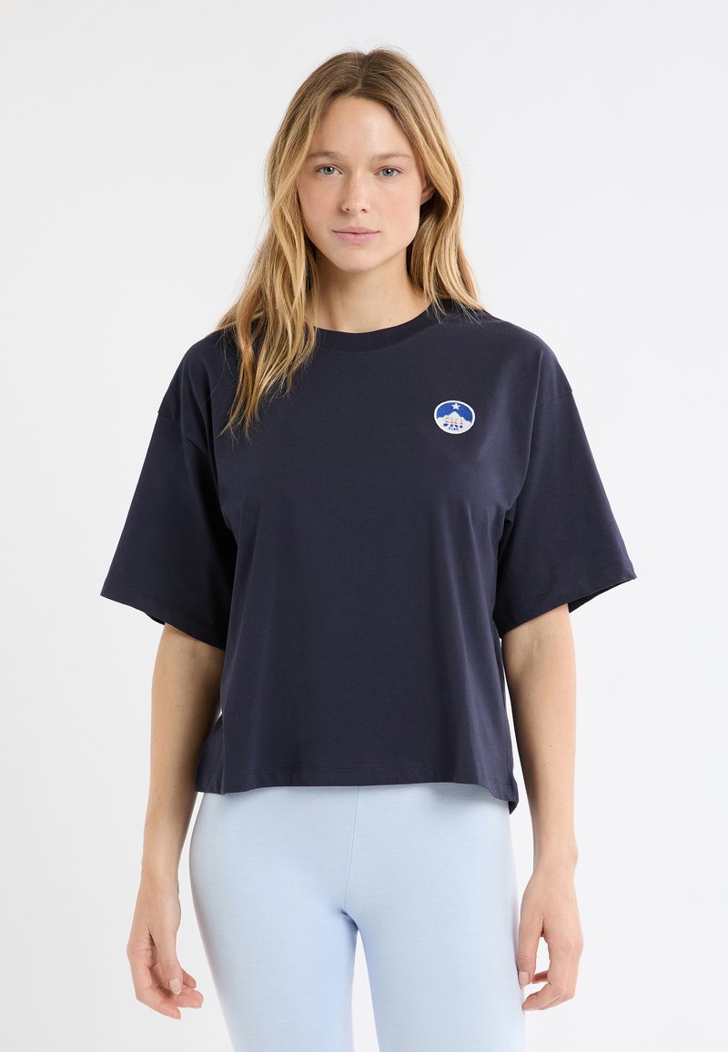 T-shirt en coton bleu marine avec un col rond et une coupe ample. Il comporte des manches courtes et un logo brodé circulaire sur le côté gauche de la poitrine.